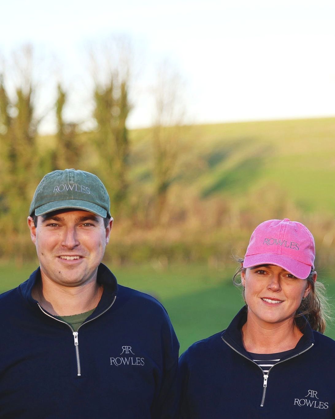 Two people wearing navy blue Rowles jackets with pink and green caps in a grassy outdoor setting.
