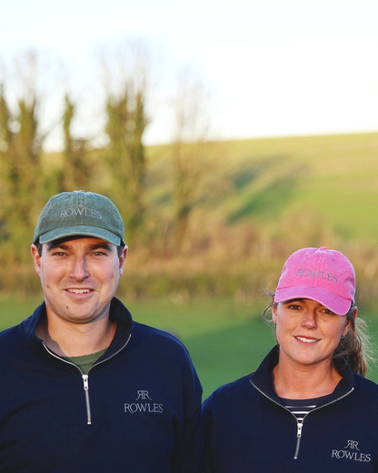 Two people wearing navy blue Rowles jackets with pink and green caps in a grassy outdoor setting.