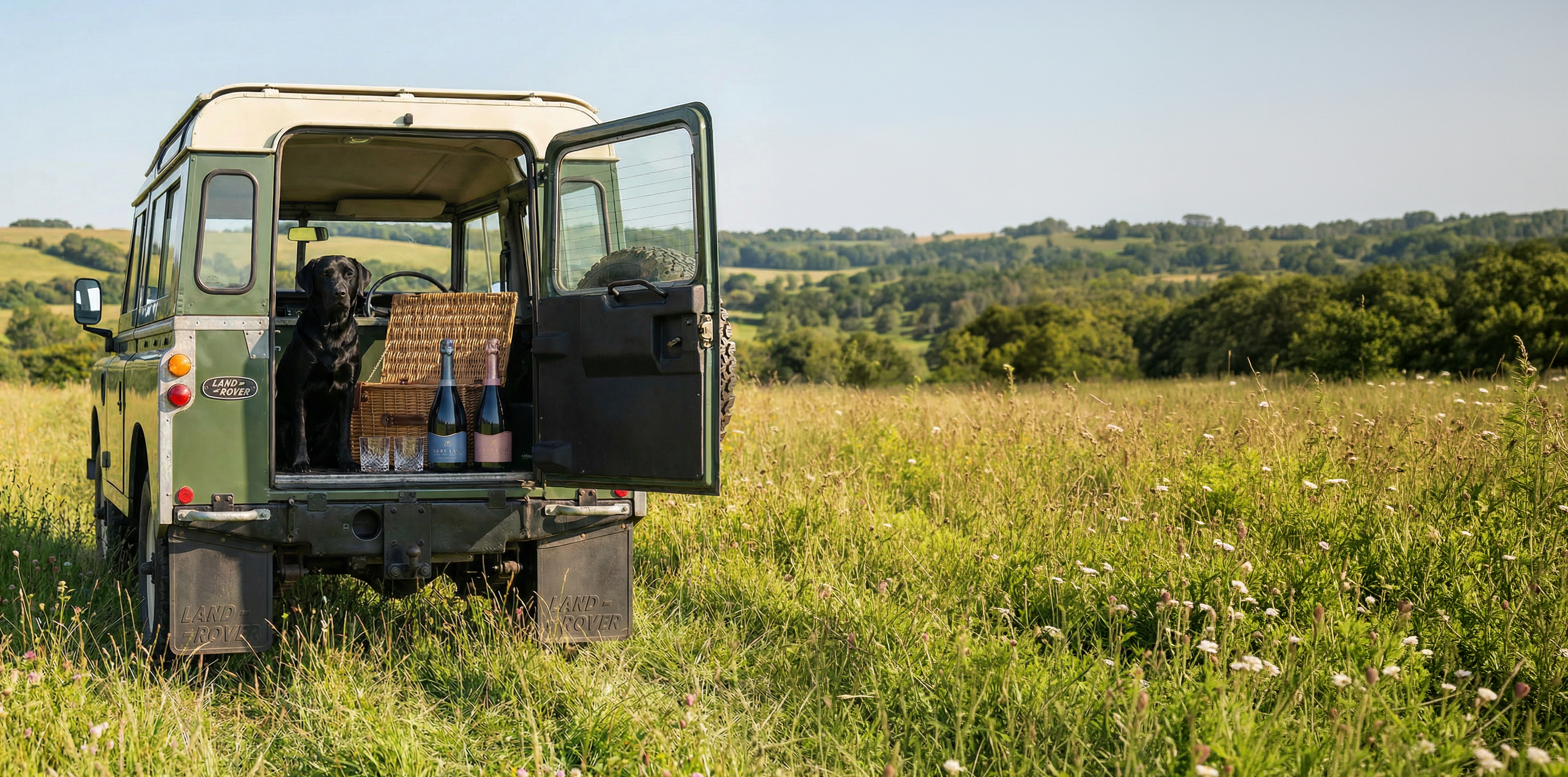 Vintage Land Rover in a grassy field with open doors and wine bottles.