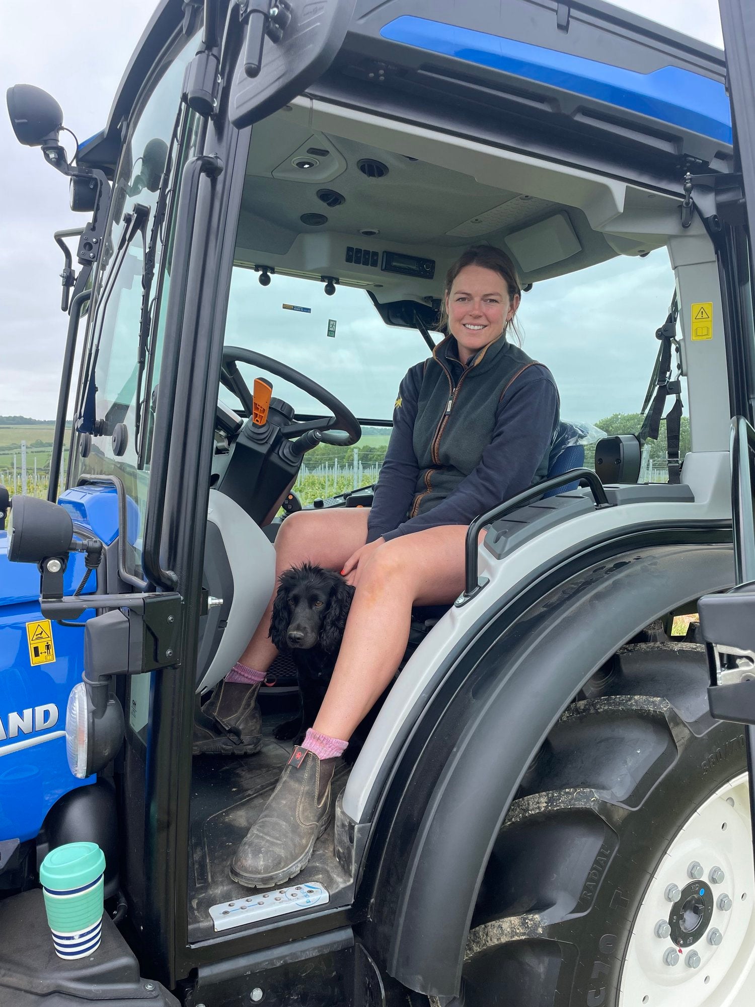 Woman sitting inside a tractor with a dog on her lap, outdoors.