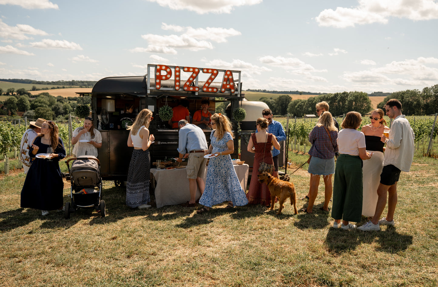 People gathered around a pizza truck in an open field on a sunny day.