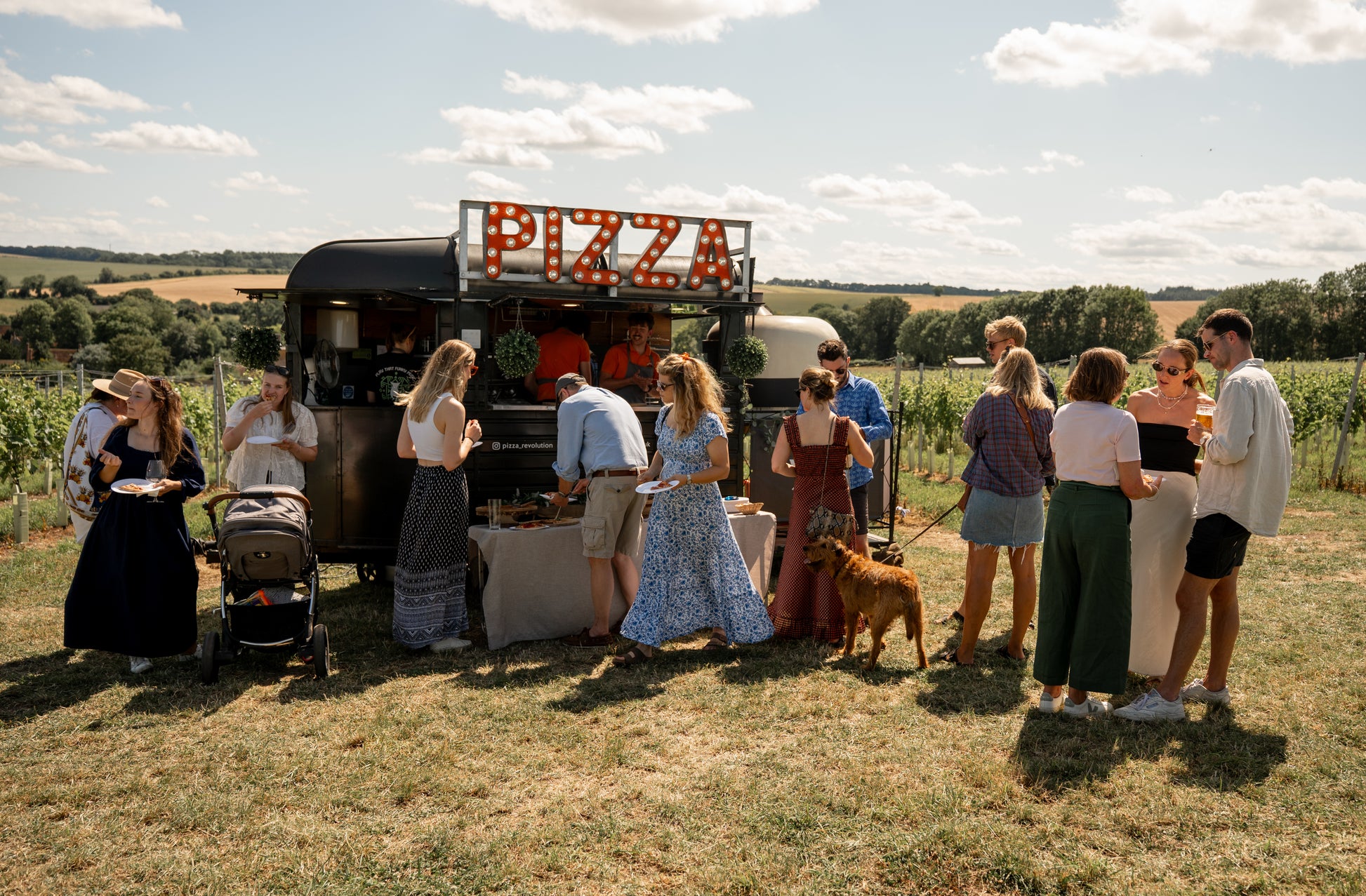 People gathered around a pizza truck in an open field on a sunny day.