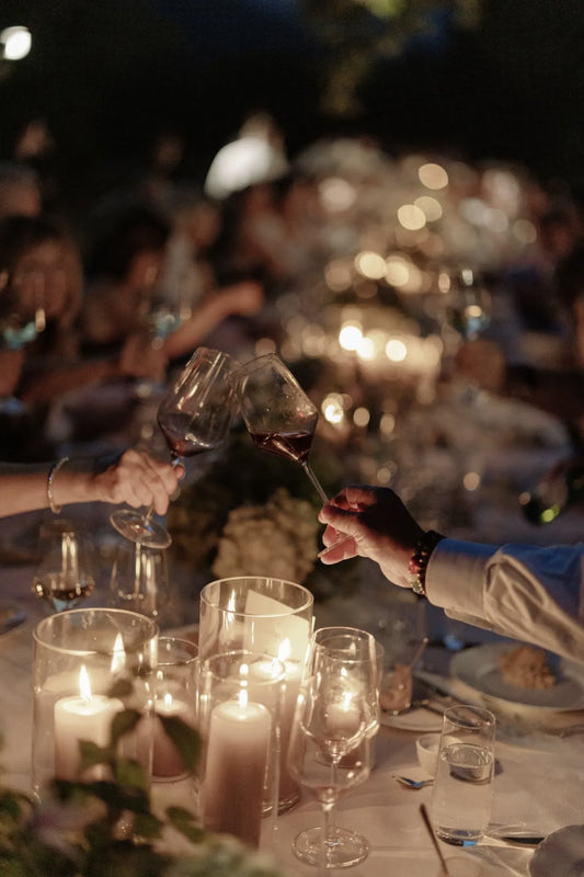Two people clinking wine glasses at a dinner table with candles and other guests in the background.
